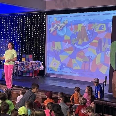 A woman in bright pink pants and a neon yellow shirt speaking to a group of children seated on the floor during a presentation or event, with a large screen displaying colorful graphics and the logo "Twist and Turns" in the background.