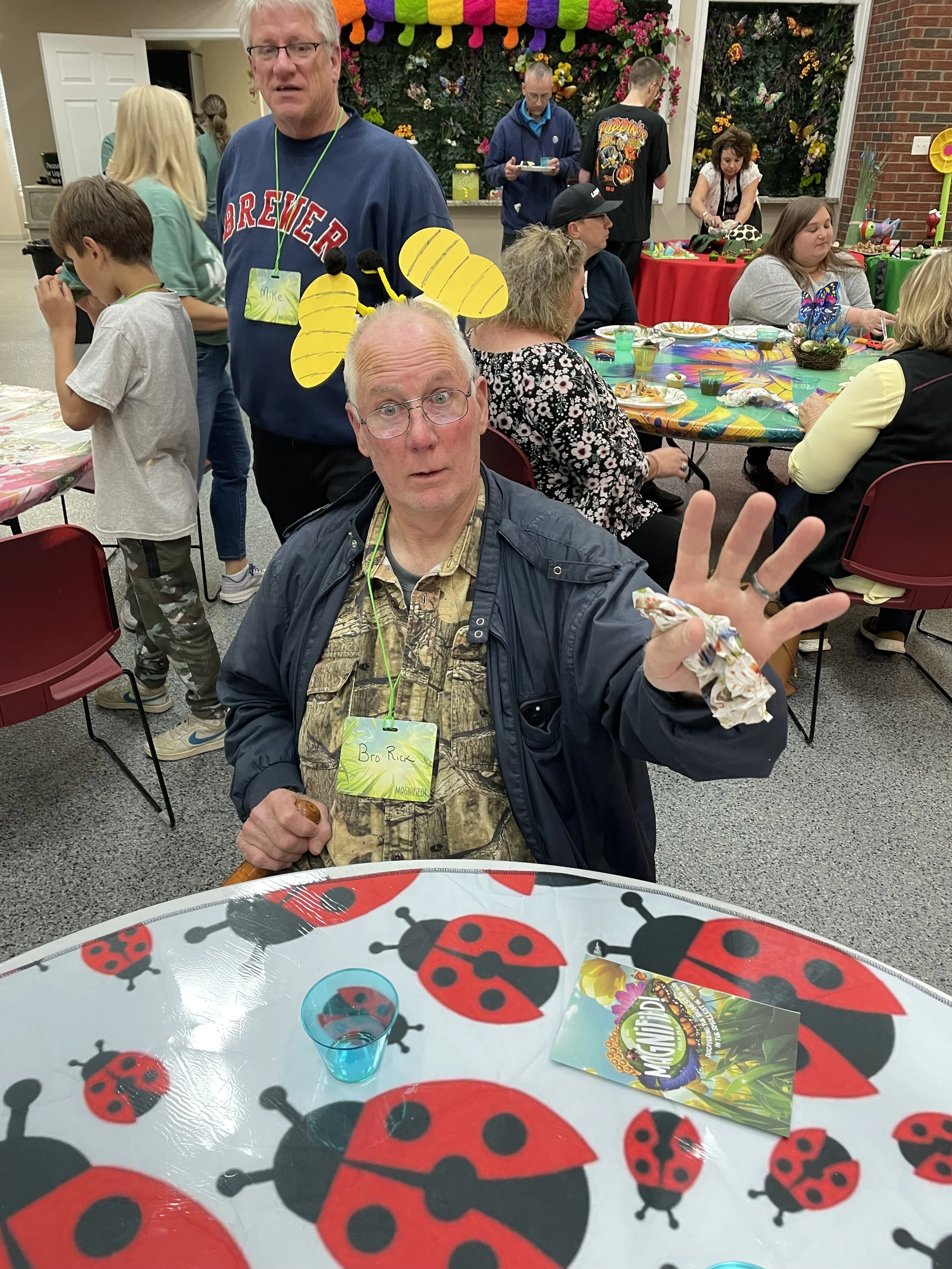 People at a party with colorful decorations and a ladybug themed tablecloth, including an older man wearing a bee headband and a younger man with a 'Brewery' shirt and a name tag that says 'Mike', in a lively indoor setting.
