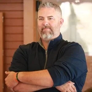 A middle-aged man with gray hair and a beard, wearing a black zip-up hoodie, standing with arms crossed in front of a window and wooden wall, outdoors.