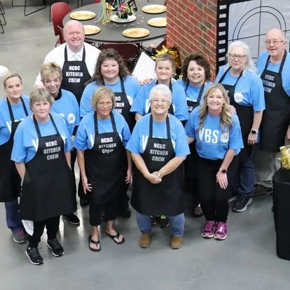 Group of people wearing blue shirts and black aprons with 'NCBC Kitchen Crew' written on them, posing for a photo indoors, possibly at a community event or gathering.