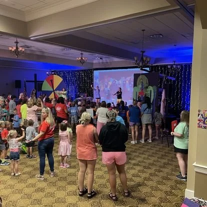 People gathering at an indoor event with a stage, large screen, colorful wheel, and blue lighting.