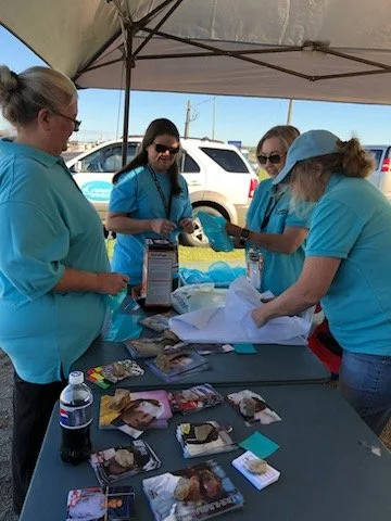 Four women in teal shirts under a canopy at an outdoor event, arranging small items and pamphlets on a table with a partially opened package, a water bottle, and photographs.