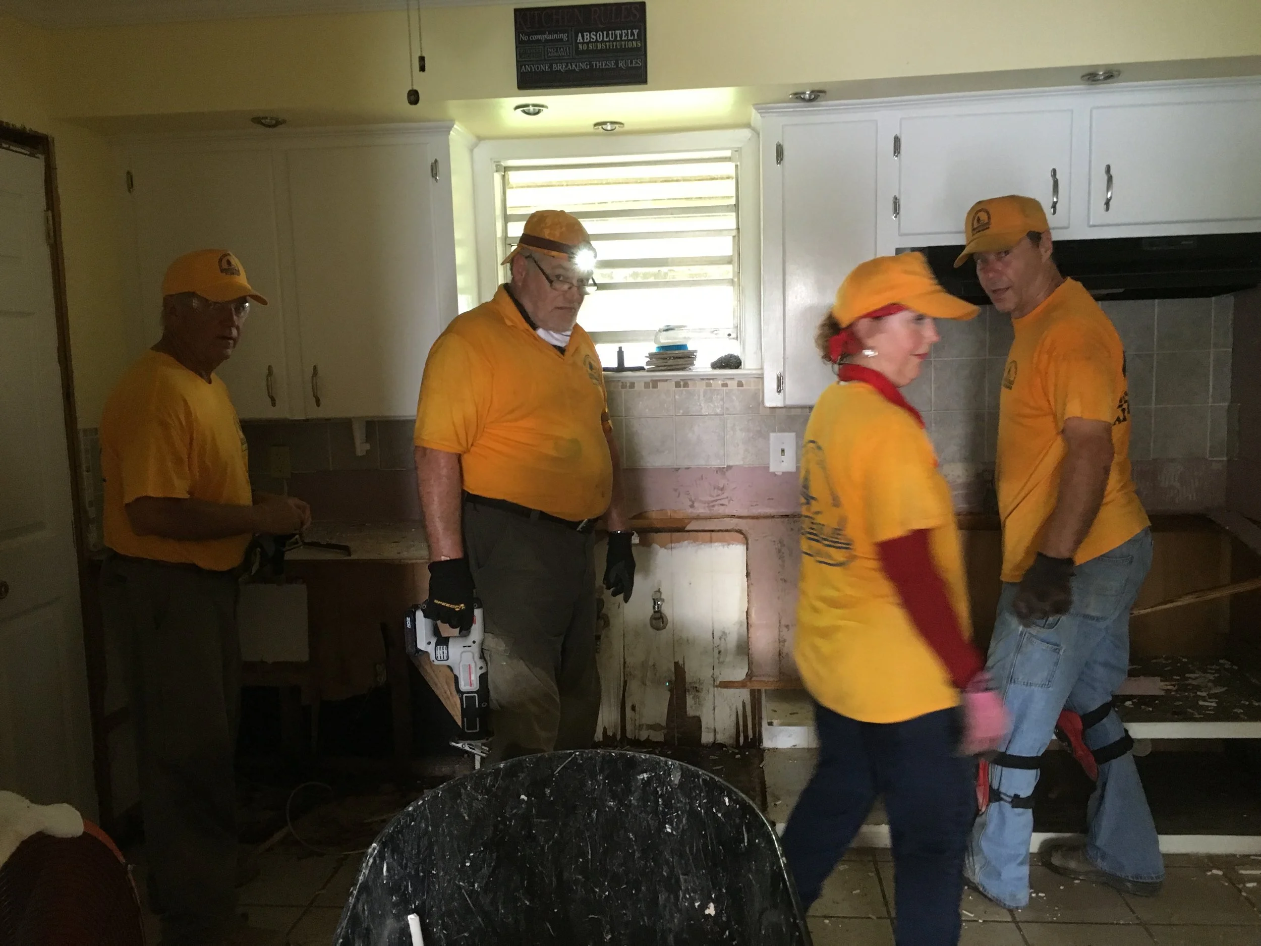 Five people wearing yellow shirts and caps in a kitchen, with one person holding a drill, appearing to clean or repair water damage; one person is blurred in motion.