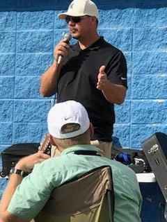 Man speaking into a microphone outdoors, wearing a black polo shirt and white cap, with a blue wall in the background.