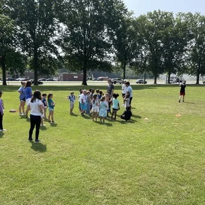 A group of children and adults gathered outdoors on a grassy field during daytime, possibly for a game or activity.