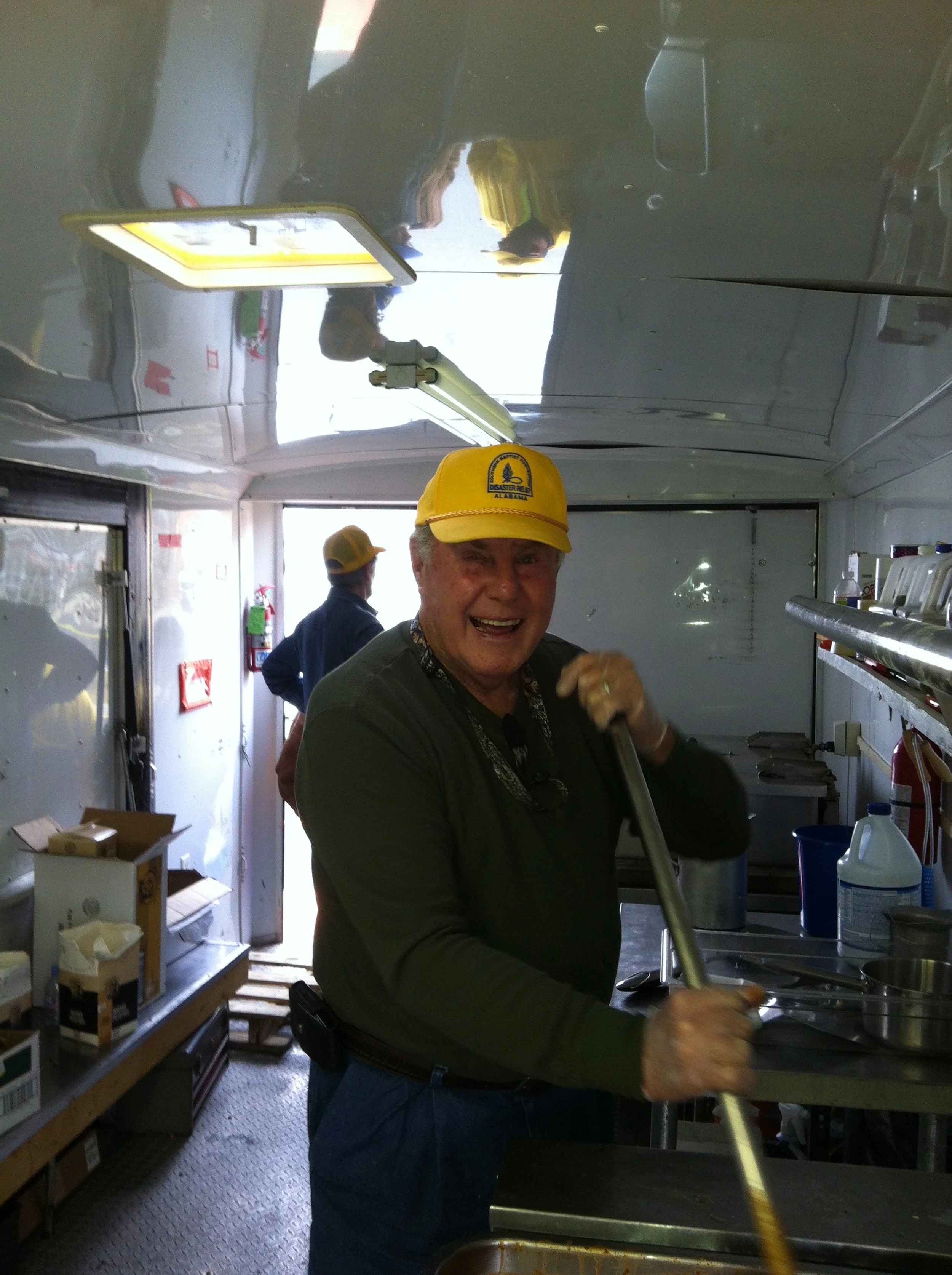 A smiling older man wearing a yellow baseball cap and dark green shirt, stirring food in a large container inside a food truck or kitchen, with two other men in yellow hats in the background.