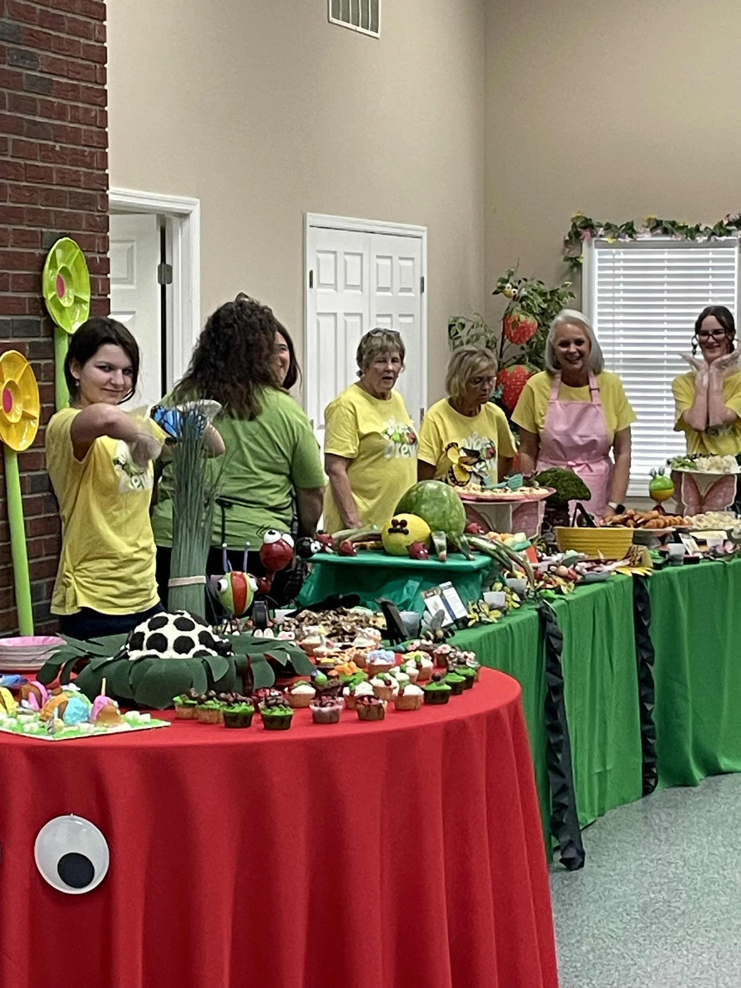 Group of women at a decorated table with cupcakes and treats, celebrating a festive event, wearing yellow shirts and aprons, with colorful decorations and themed display.