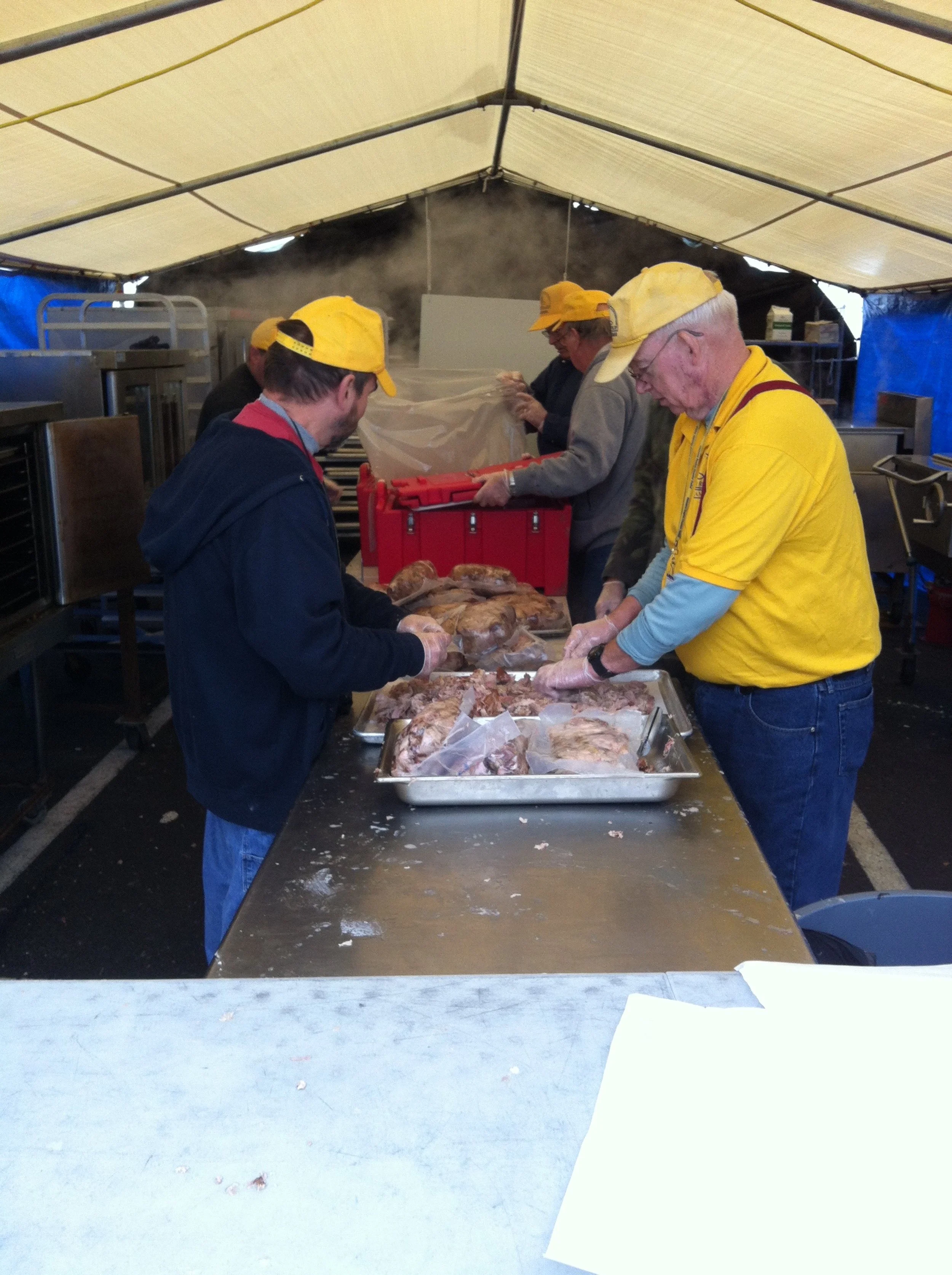People working at a food preparation station, carving and preparing large pieces of meat, under a tent with yellow hats and gloves.