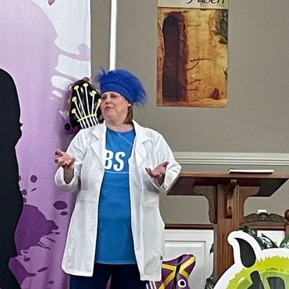 Woman speaking at a presentation or event, wearing a blue headwrap and a white lab coat, standing in front of a purple backdrop with abstract designs.