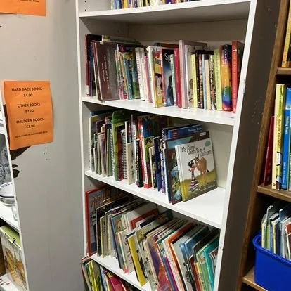 A white bookshelf filled with children's books in a library or bookstore. There are four shelves with colorful book covers, and a bright orange sign describing book prices on the side.