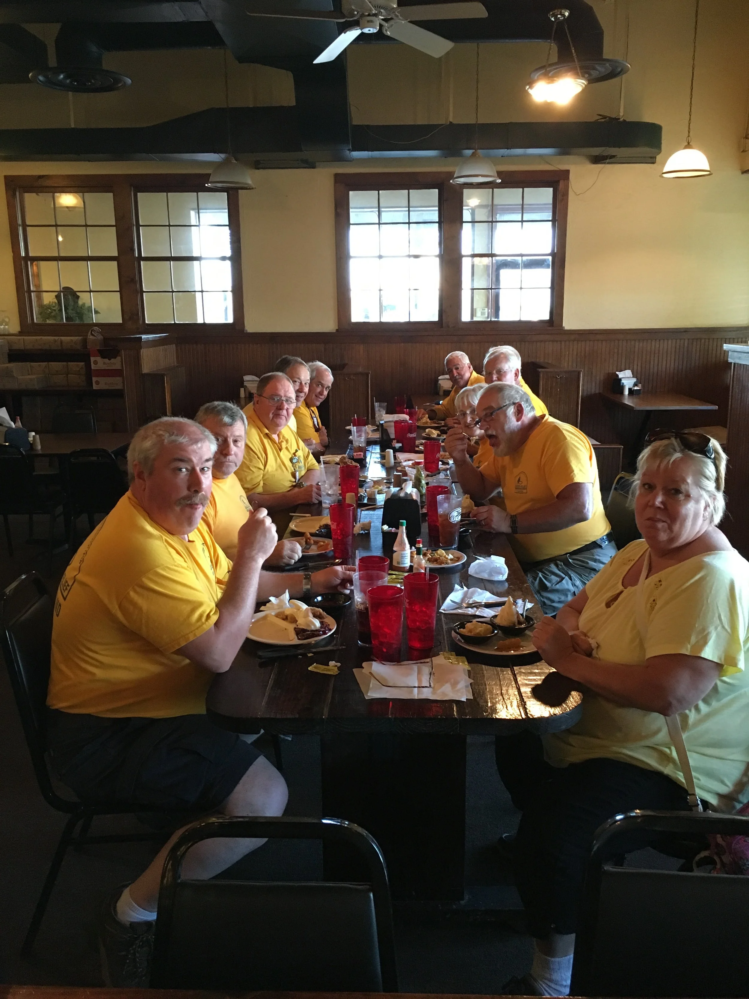 A group of eleven people wearing yellow shirts sitting around a long restaurant table, enjoying a meal, with plates of food, cups, and condiments visible.