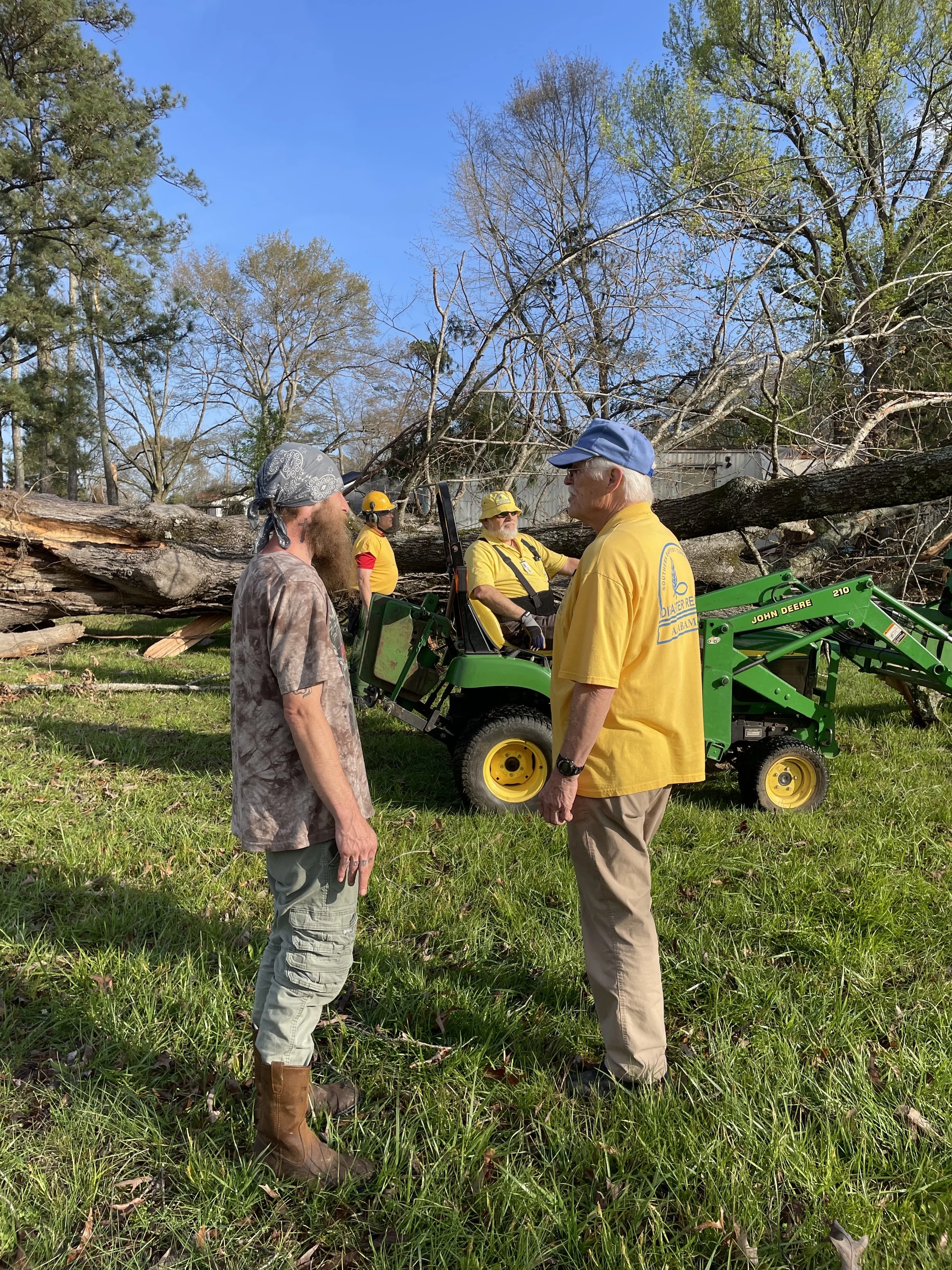 Four men, two in yellow shirts and helmets, one in a gray bandana, and one in a yellow shirt, standing outdoors in front of fallen trees; two men are seated on a green John Deere compact tractor, with trees and a blue sky in the background.