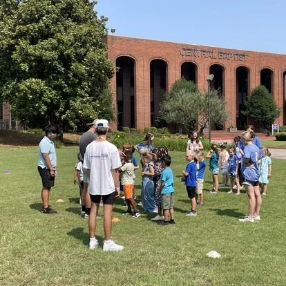 Children and adults gathered outside the Central Baptist church building on a grassy lawn.