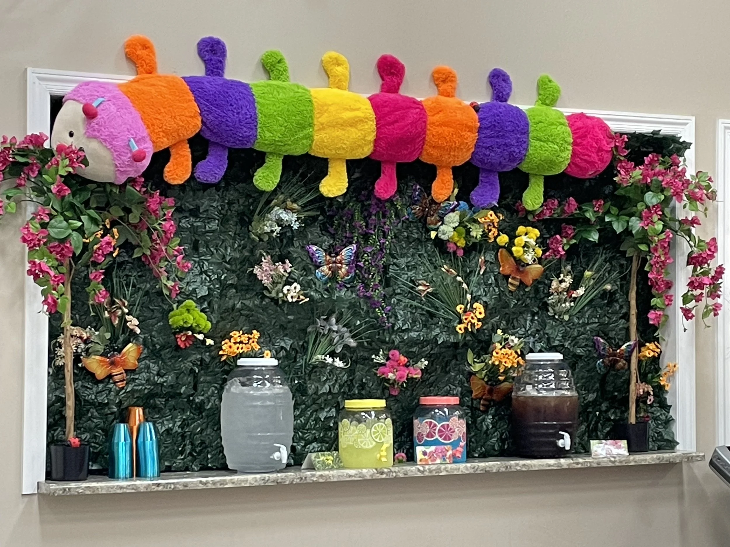 Colorful caterpillar plush toy hanging above a floral display with butterflies and flower decorations, set on a marble countertop with jars of drinks and cups.