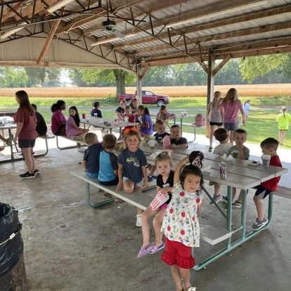 Children seated at picnic tables under a pavilion, with adults and a red truck in the background, at an outdoor gathering.