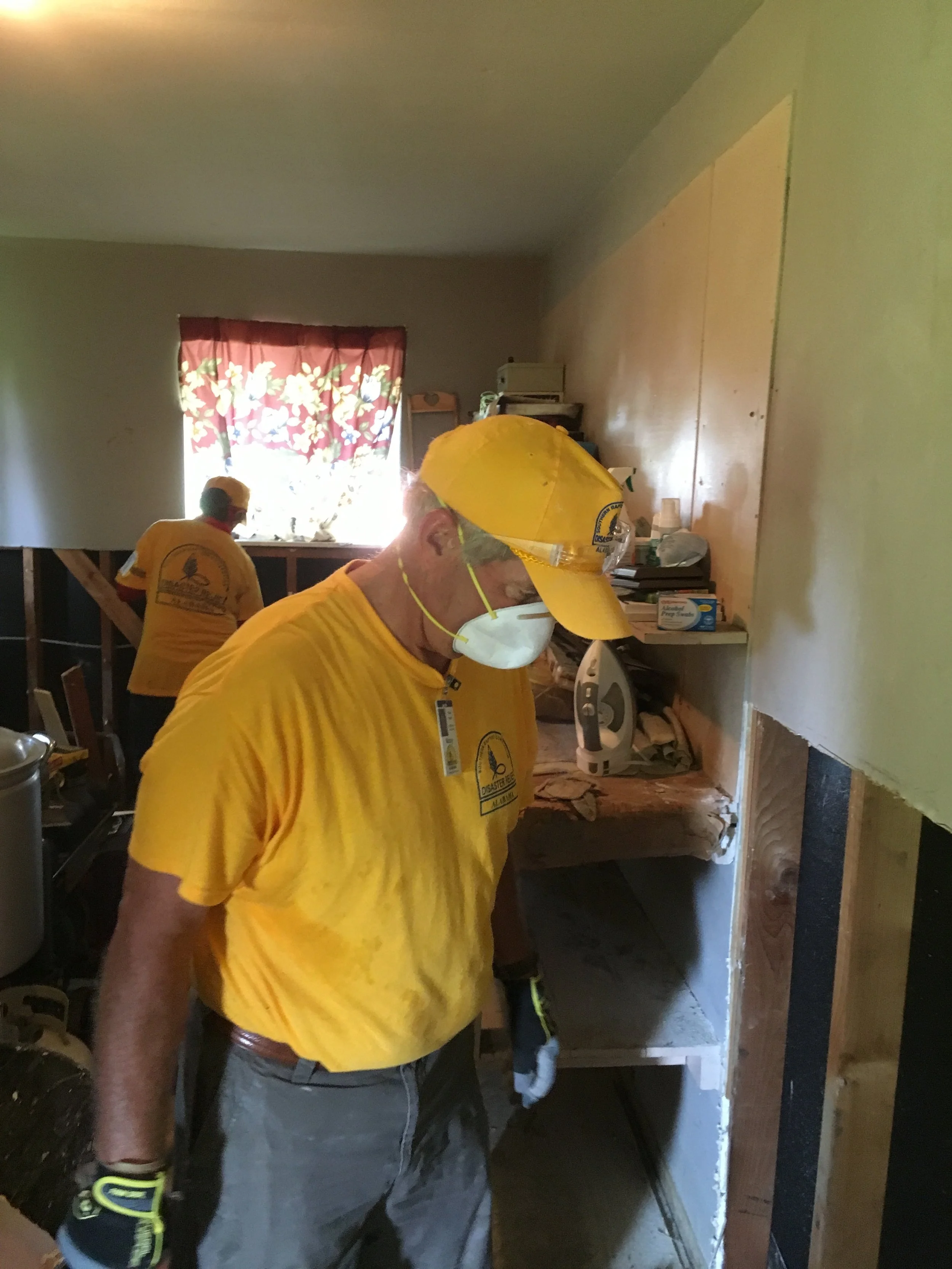 Construction worker in yellow shirt, cap, mask, and gloves working inside a house with exposed wall framing and a shelf with tools.