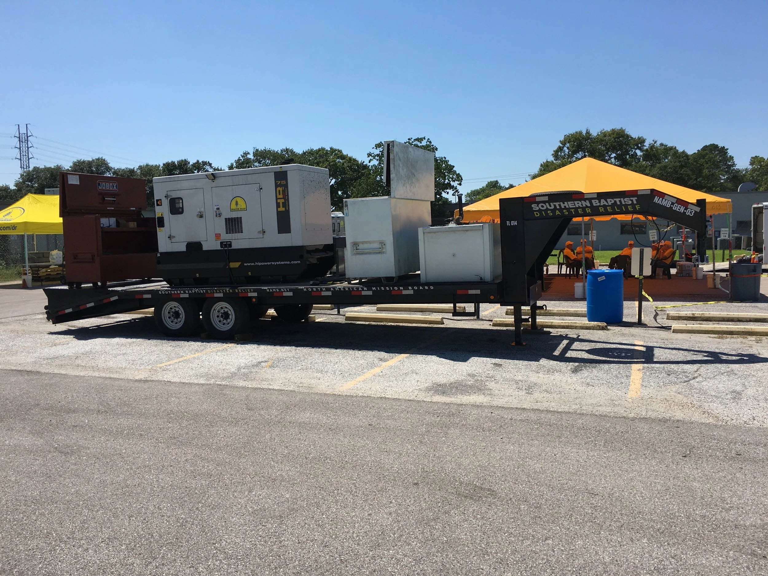 A mobile generator and storage units on a trailer parked in a parking lot with a yellow tent and people sitting under an orange tent in the background.