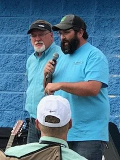 Two men speaking into a microphone at an outdoor event, with an audience member wearing a white cap in the foreground, against a blue wall background.