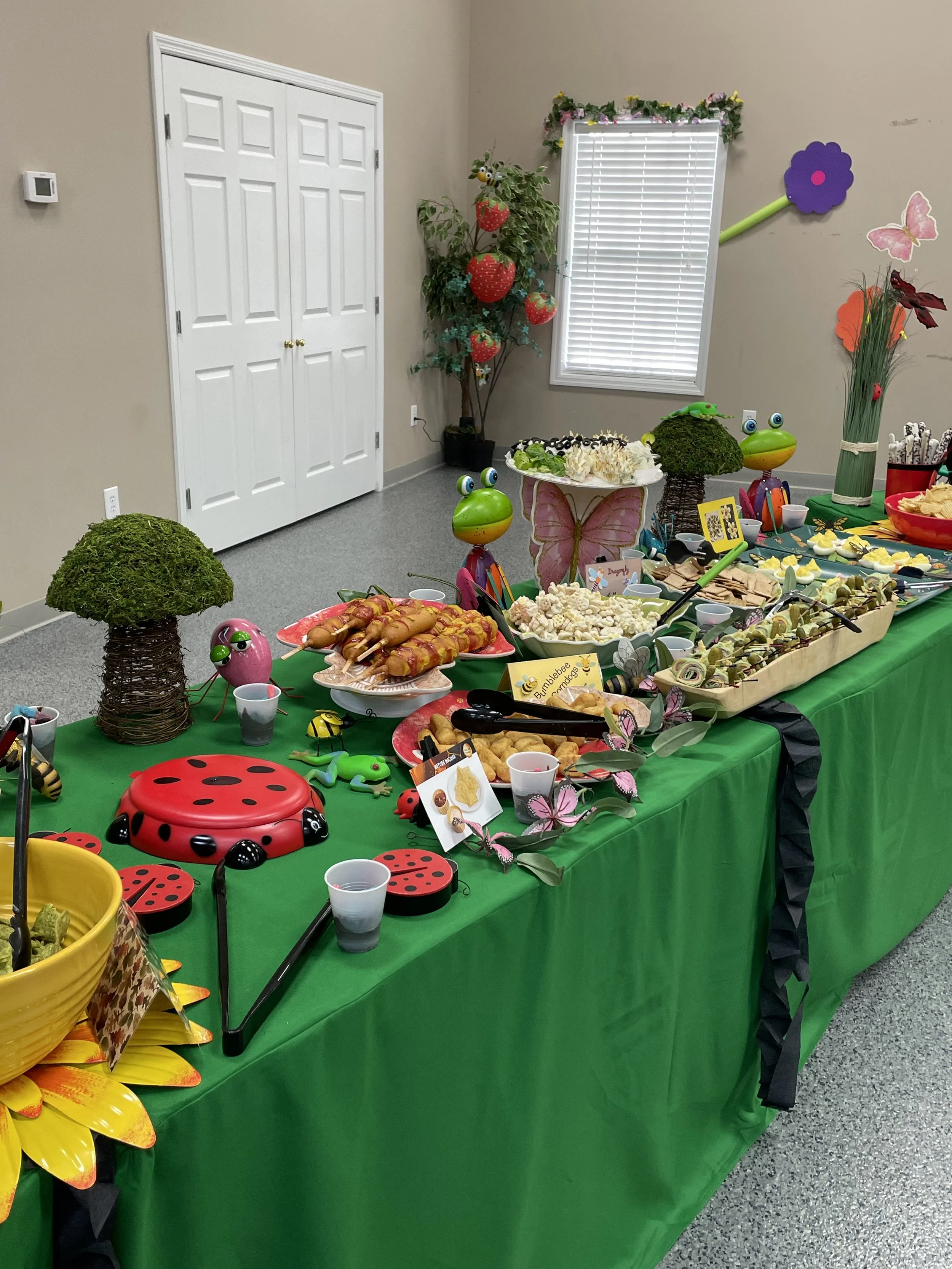 A dessert table decorated with ladybug and butterfly themed items, featuring snacks and treats like fruit, chips, and popcorn, with a nature-inspired theme, including toys, flowers, and animal decorations, set in a room with beige walls and a white d