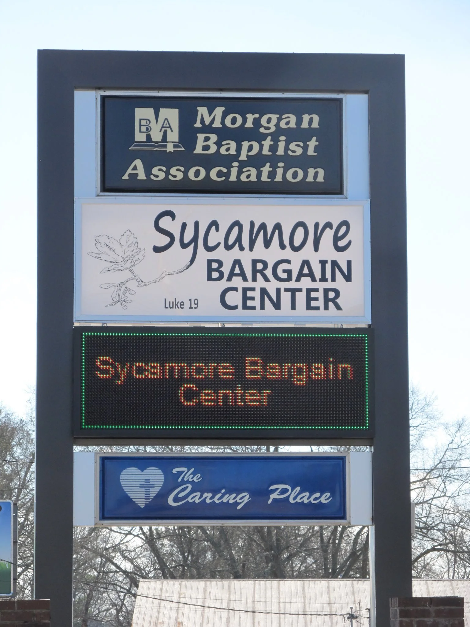 Signboard for Morgan Baptist Association and Sycamore Bargain Center, with an electronic display showing the shop's name and a blue sign reading "The Caring Place" with a heart logo.