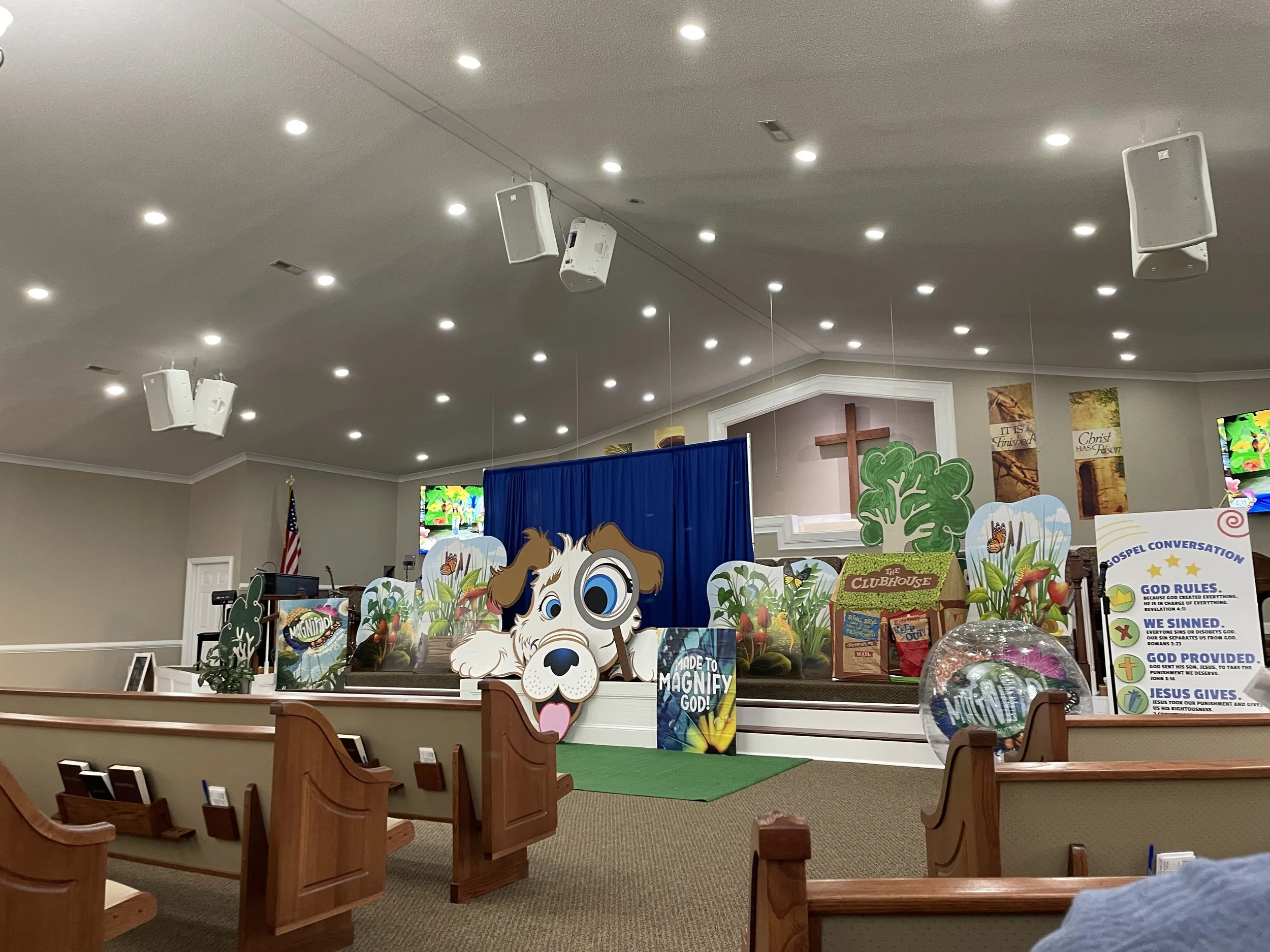 Inside a church with a decorated stage featuring cartoon-style animals, a cross, and banners with religious messages, prepared for a children's event or Sunday school.