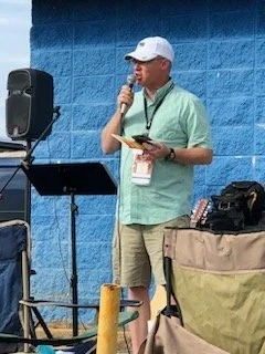 Man speaking into microphone at outdoor event, holding a booklet, with sound equipment and a blue wall in the background.