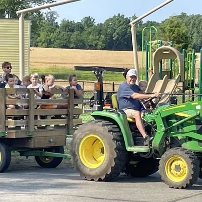 Child driving a green tractor with a trailer of children riding behind.