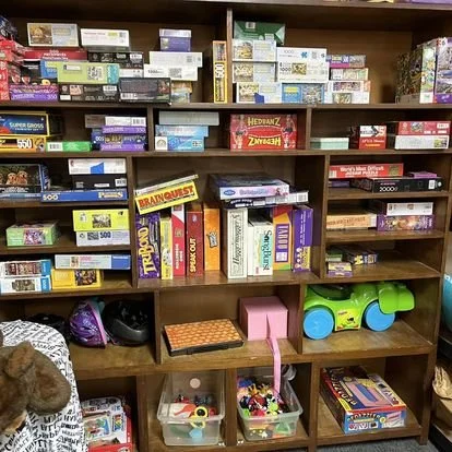 Shelves filled with board games, puzzles, and toys, including a Green and Blue toy truck, a pink item, and various colorful game boxes.