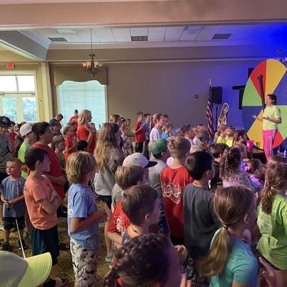 A large group of children gathered in a spacious room, facing a woman in a yellow shirt next to a colorful spinning wheel, with American and other flags in the background.