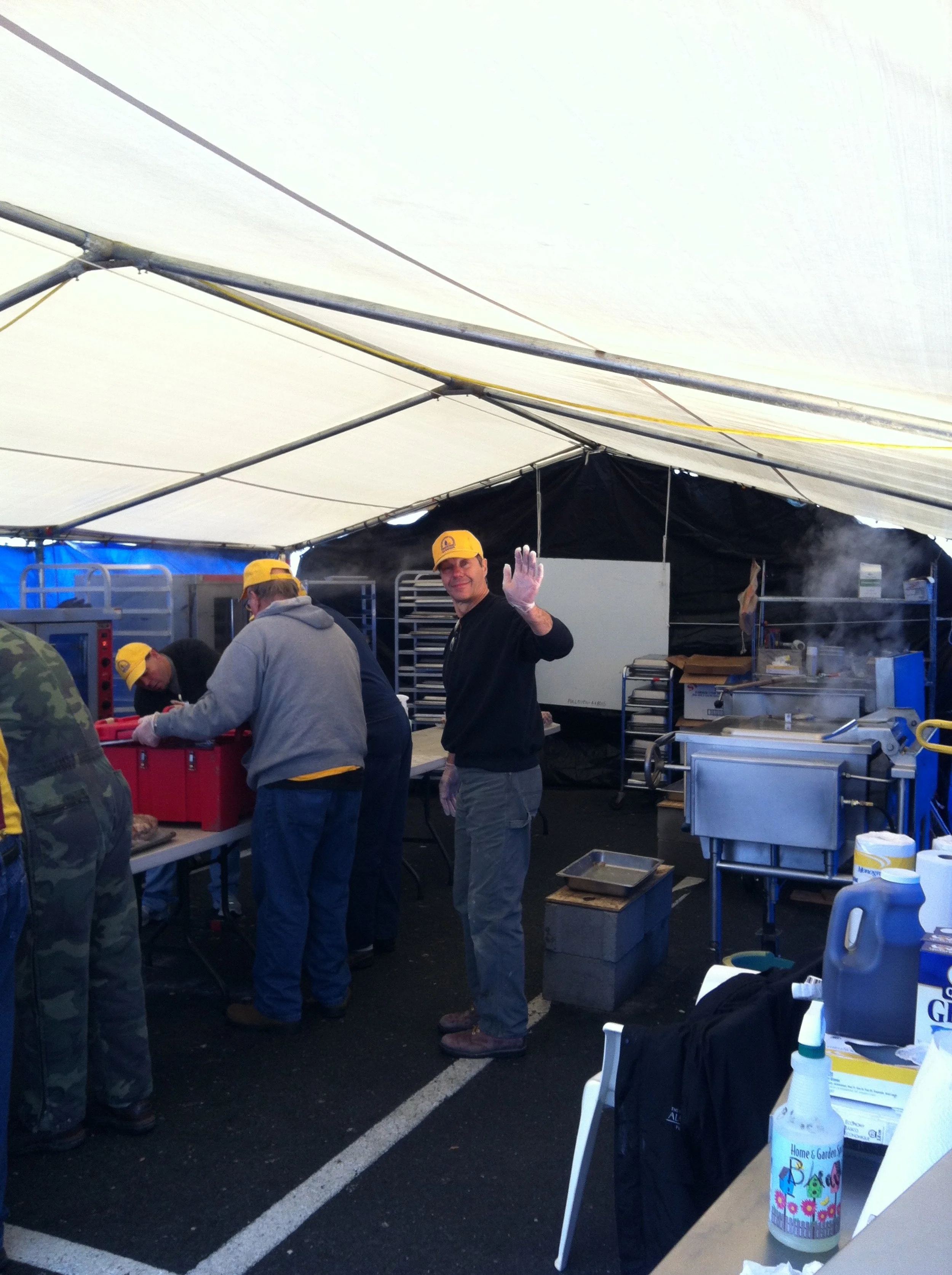 Group of people working under a large white tent, wearing yellow hats and gloves, with a man smiling and waving at the camera, surrounded by cooking and kitchen equipment, including a steam cooker, trays, and cleaning supplies.