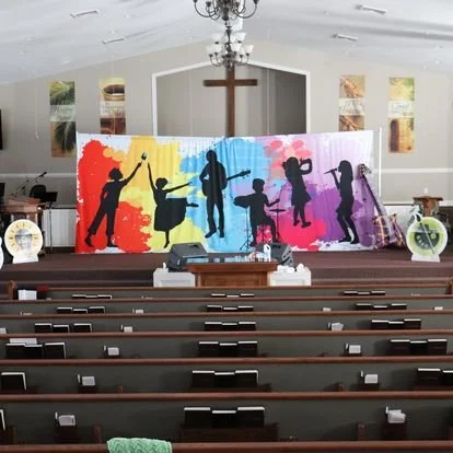 Interior of a church with a colorful backdrop of children silhouettes on stage, a large wooden cross on the wall, and church pews.