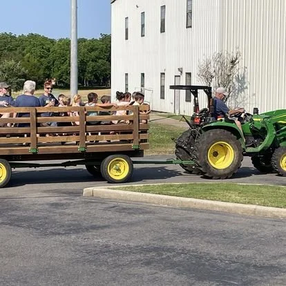 A group of people riding in a wooden wagon pulled by a green tractor on a street near a white building.