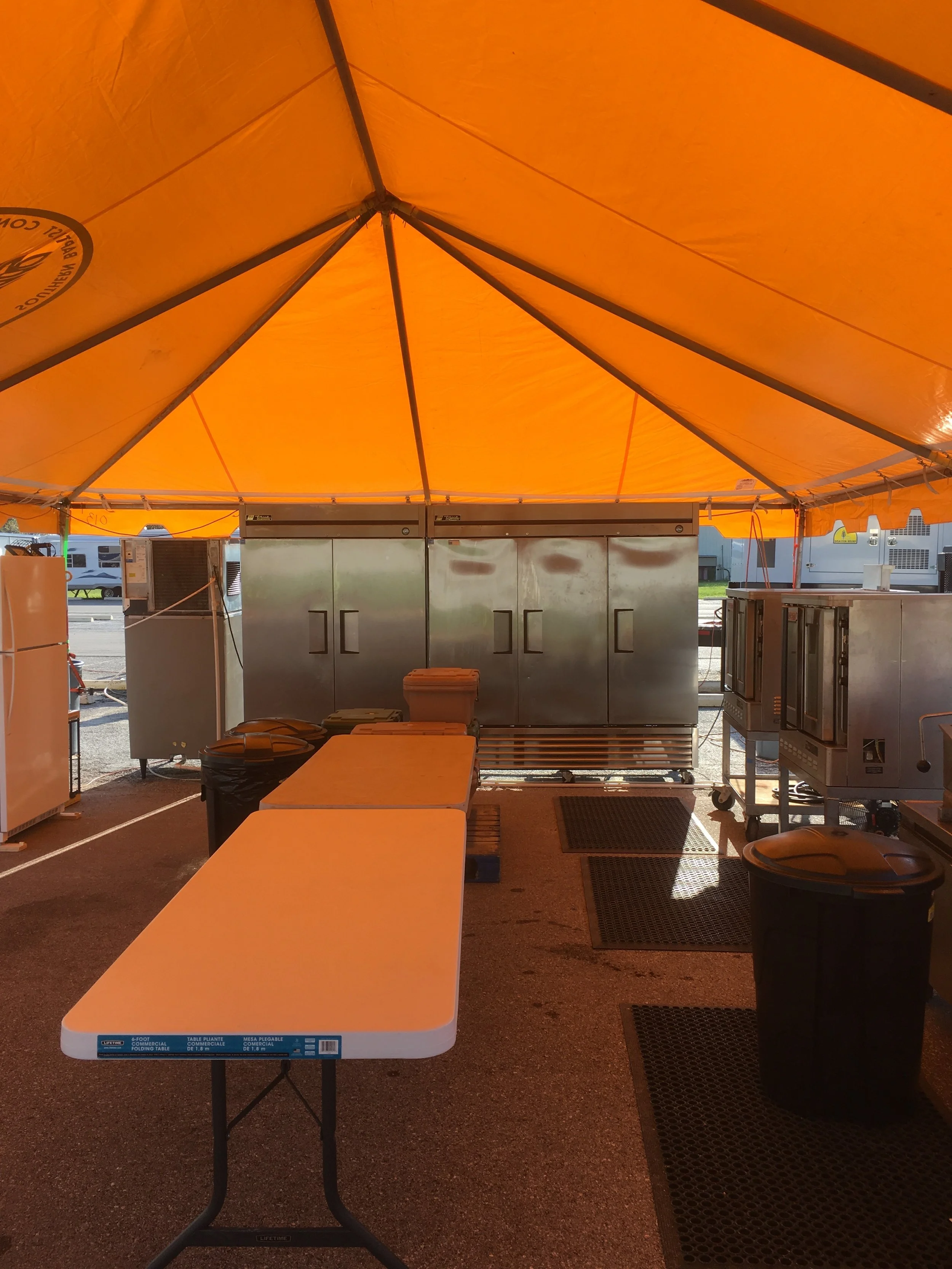 Kitchen area inside a large orange tent with stainless steel appliances, foldable tables, black trash cans, and rubber floor mats.