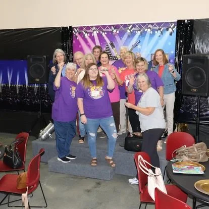 Group of women on a stage during a karaoke event, holding microphones with a colorful backdrop and sound equipment.