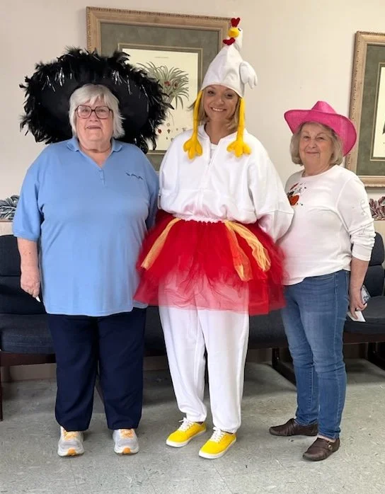 Three women standing indoors, wearing playful costumes. The woman on the left is wearing a large black feathered headpiece, glasses, a blue shirt, and dark pants. The woman in the middle is dressed as a chicken with a white hat with a red comb, a whi