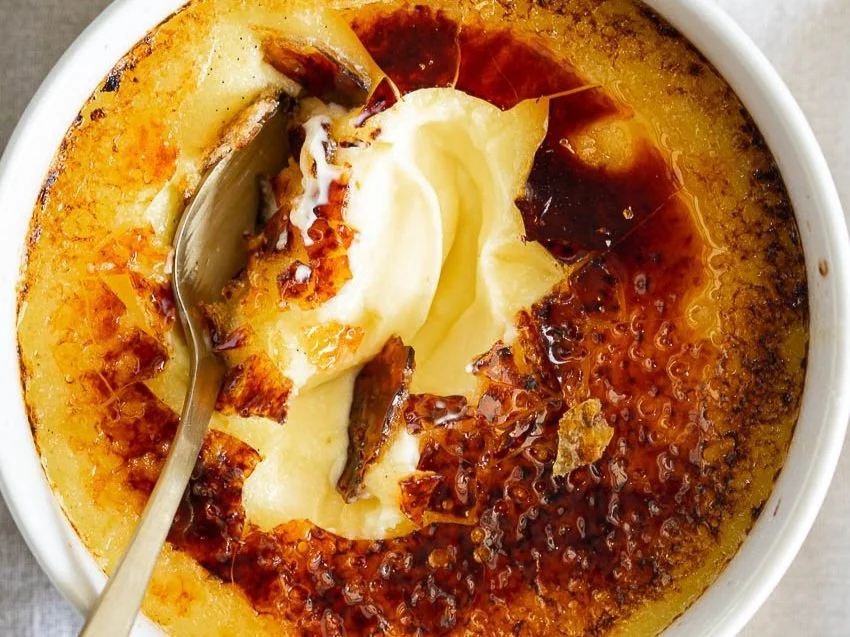 Top view of crème brûlée with a broken caramelized sugar crust, revealing creamy custard inside, and a spoon inserted into the dessert in a white ramekin.