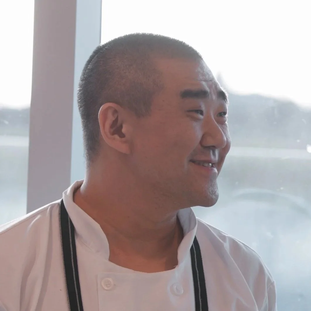 A chef in a white uniform and glasses standing in a restaurant or gallery with paintings on the walls.