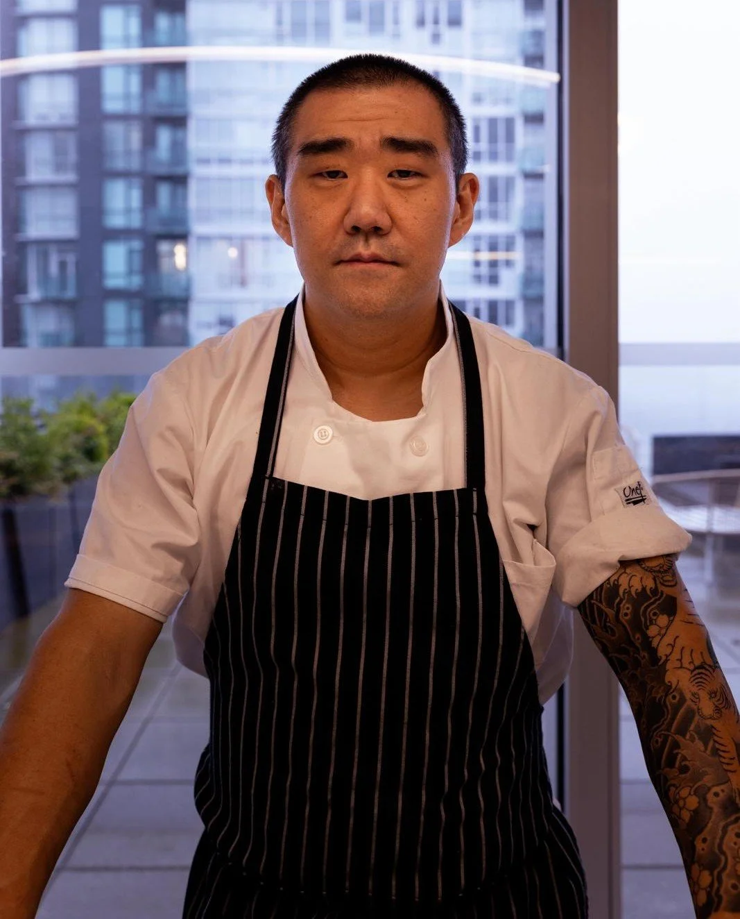A chef in a white uniform and glasses standing in a restaurant or gallery with paintings on the walls.