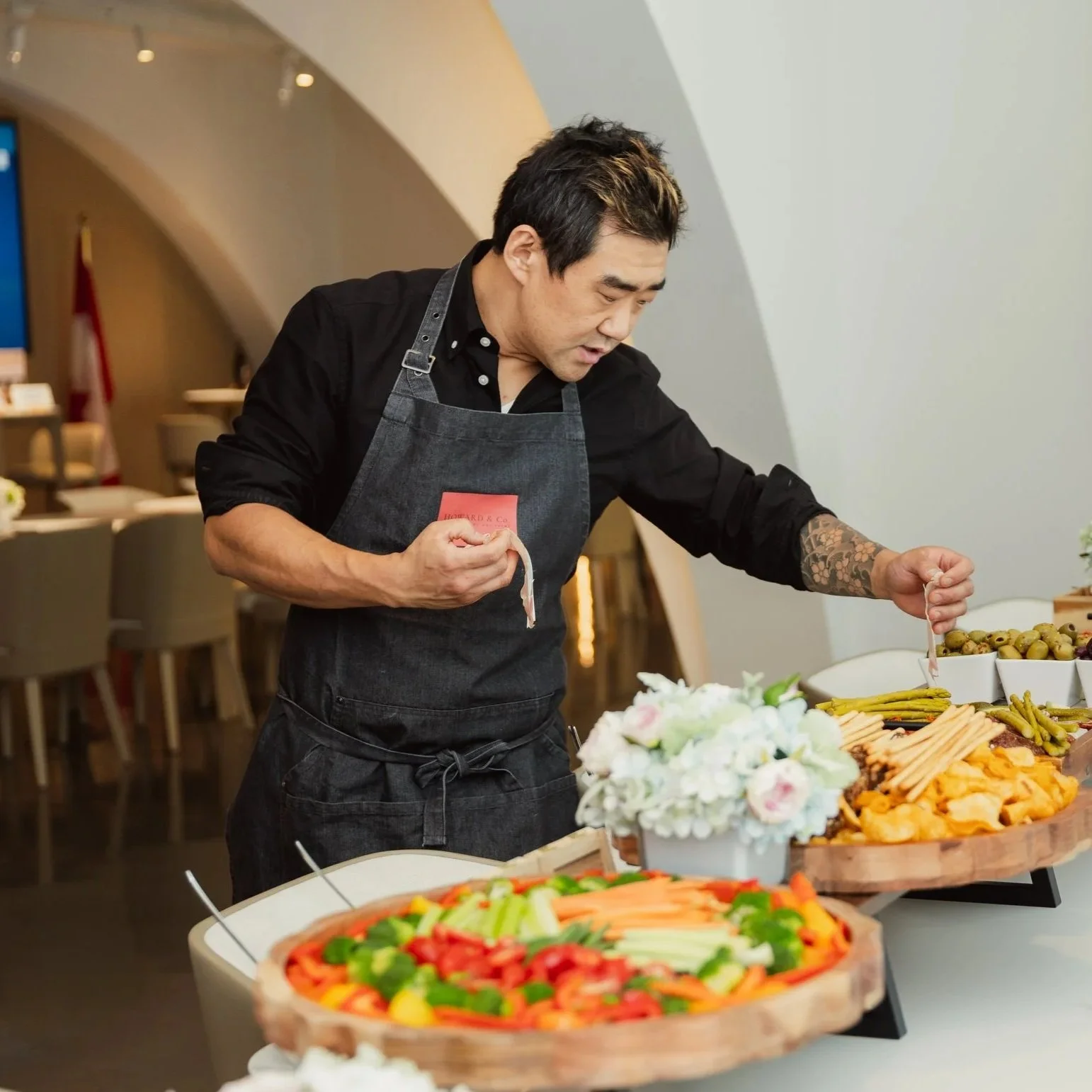 A chef in a white uniform and glasses standing in a restaurant or gallery with paintings on the walls.