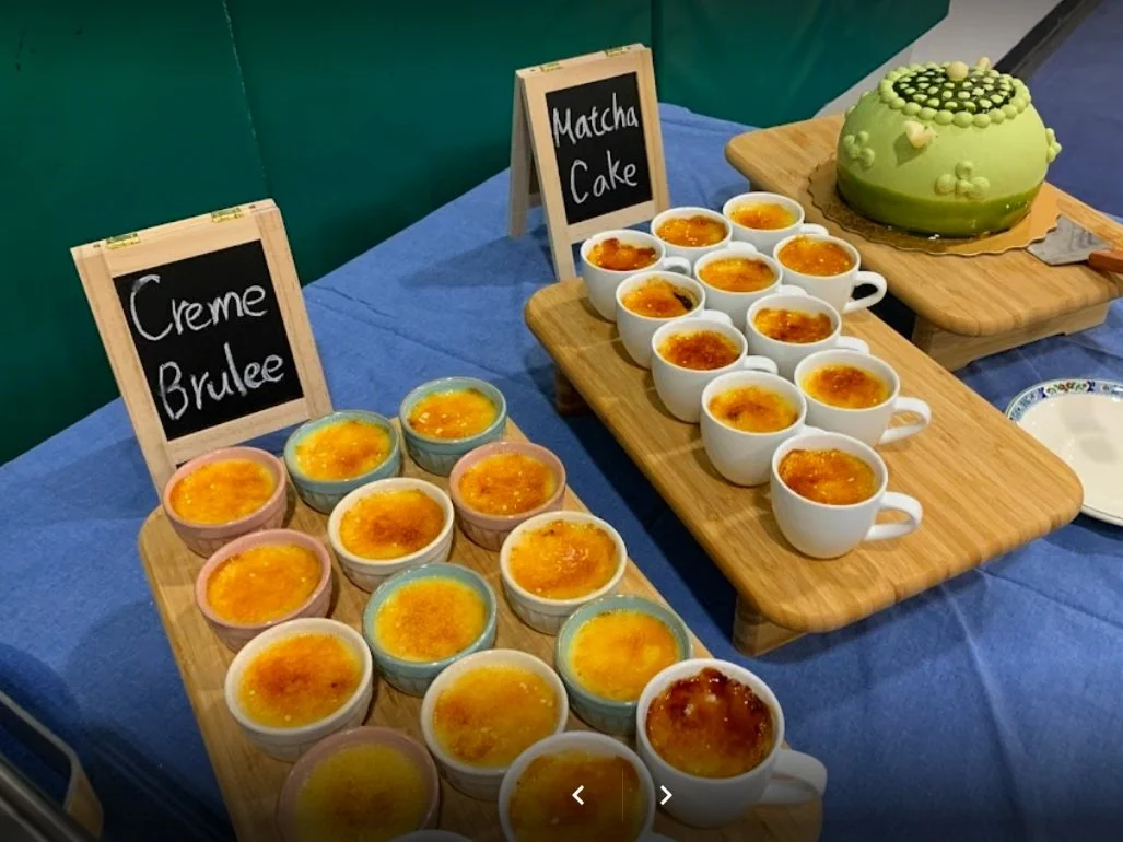 Dessert table with creme brulee in colorful ramekins, cups, and a green matcha cake on wooden boards with small signs.