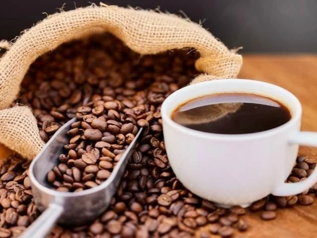Coffee beans spilling from a burlap sack with a metal scoop and a white cup of black coffee on a wooden surface.