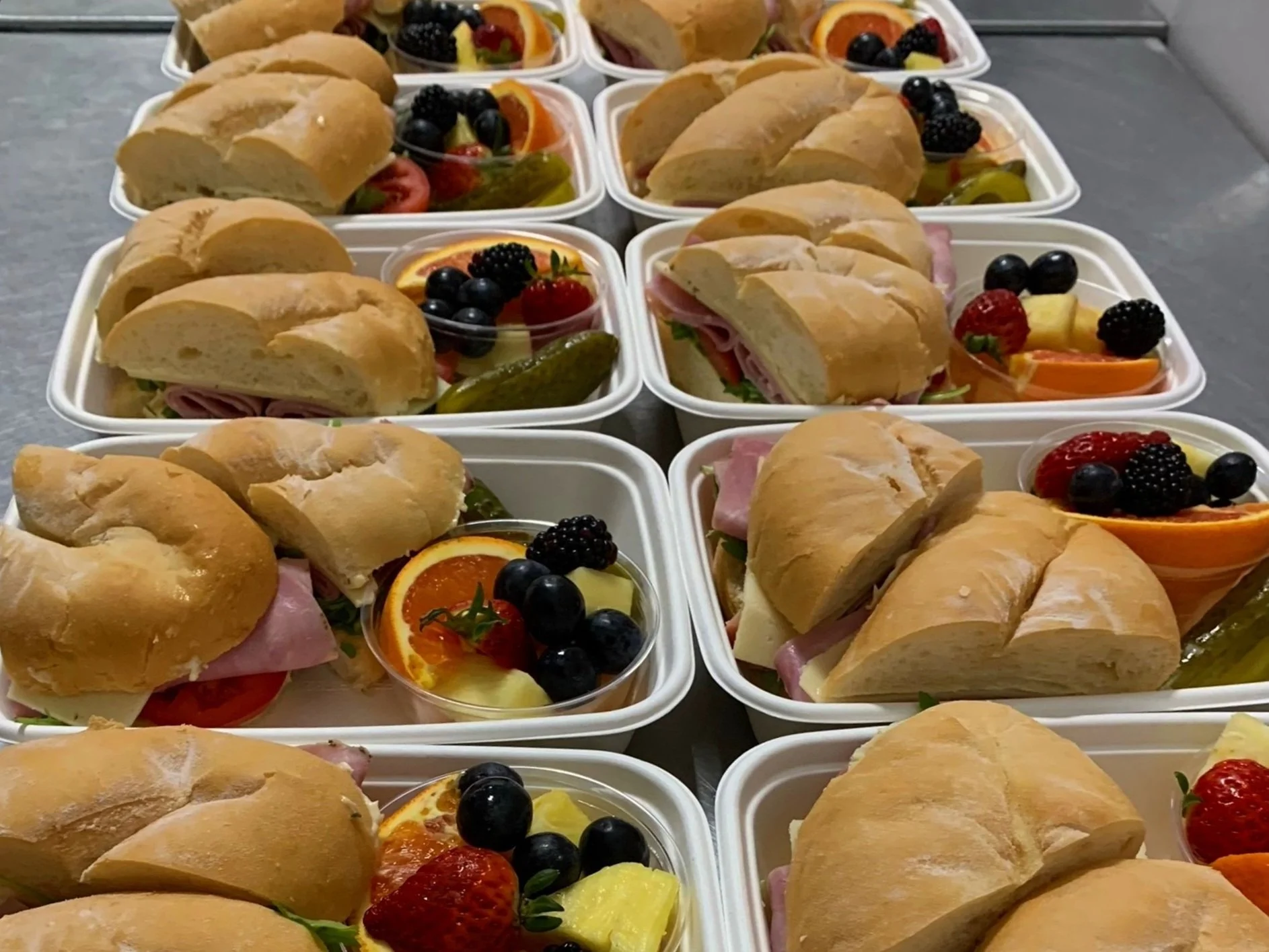 Assorted sandwich lunch boxes with ham, cheese, and lettuce, accompanied by fruit slices and pickles.