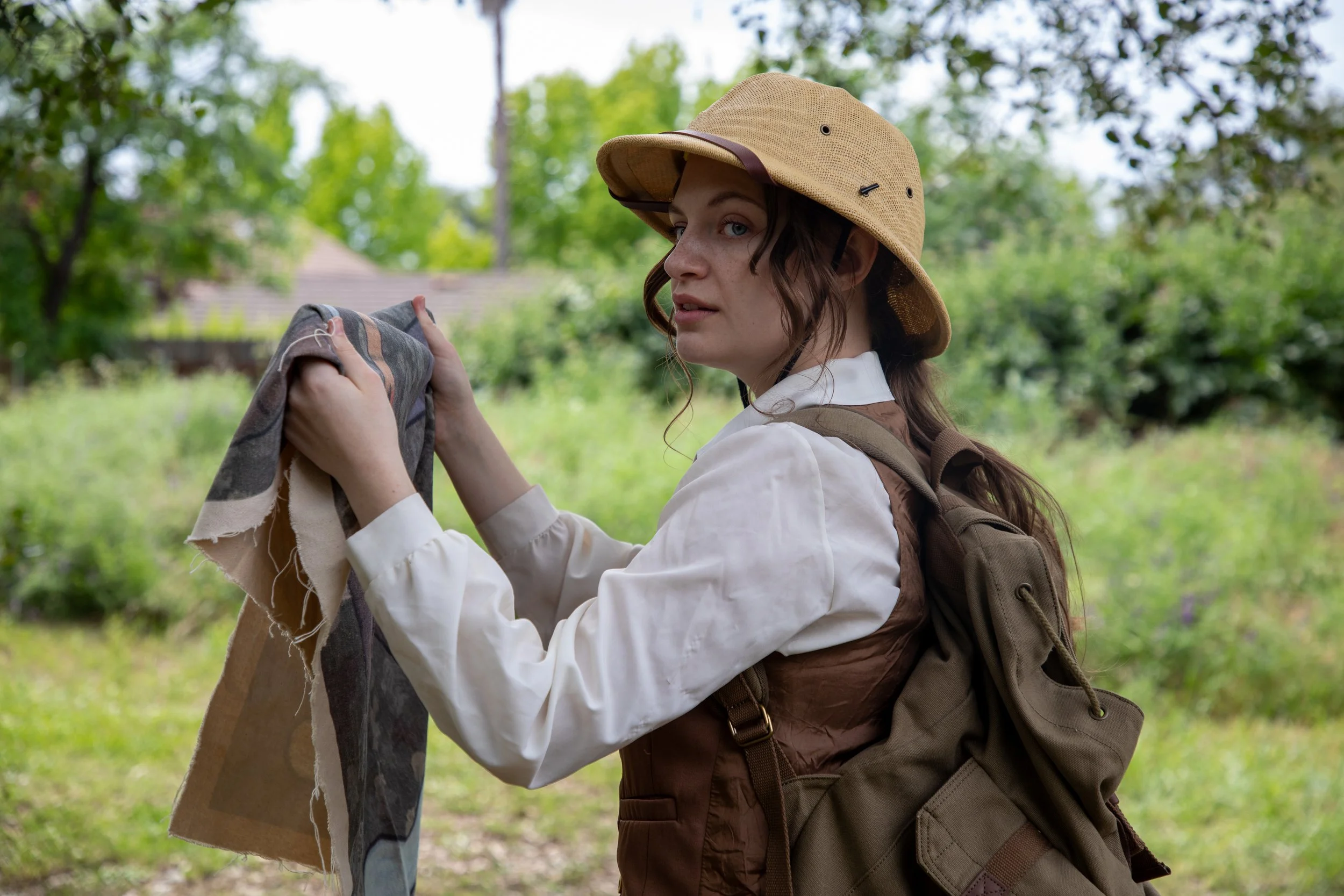 Rachel Janine Harris, wearing safari gear, a white blouse, and a brown vest is holding a map outdoors surrounded by green trees.