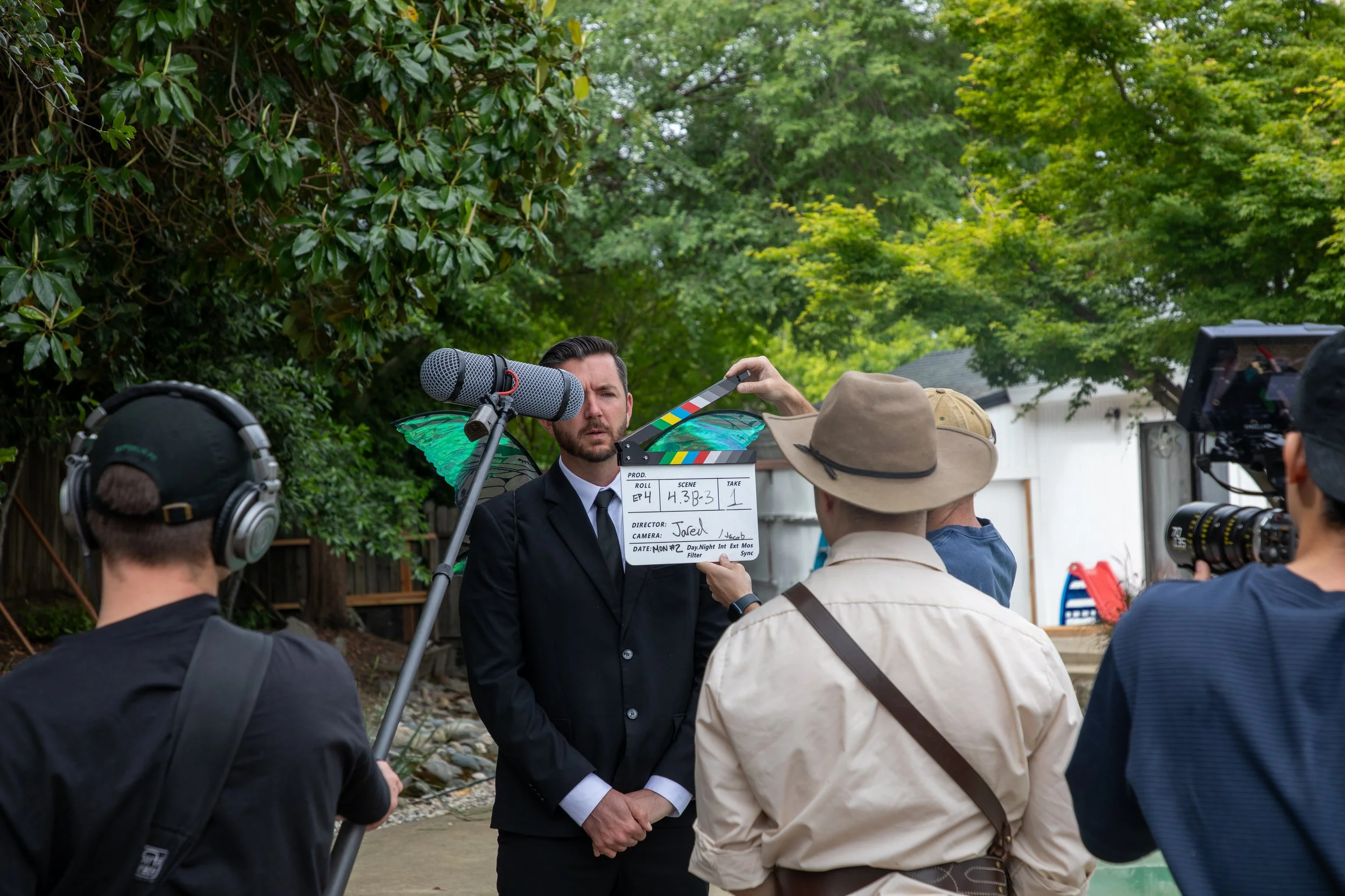 On a film set, a dressed in a black suit with green wings is being filmed while holding a clapperboard. Crew members are surrounding him, with some wearing headphones and filming equipment nearby. The scene is outdoors, with lush green trees in the b