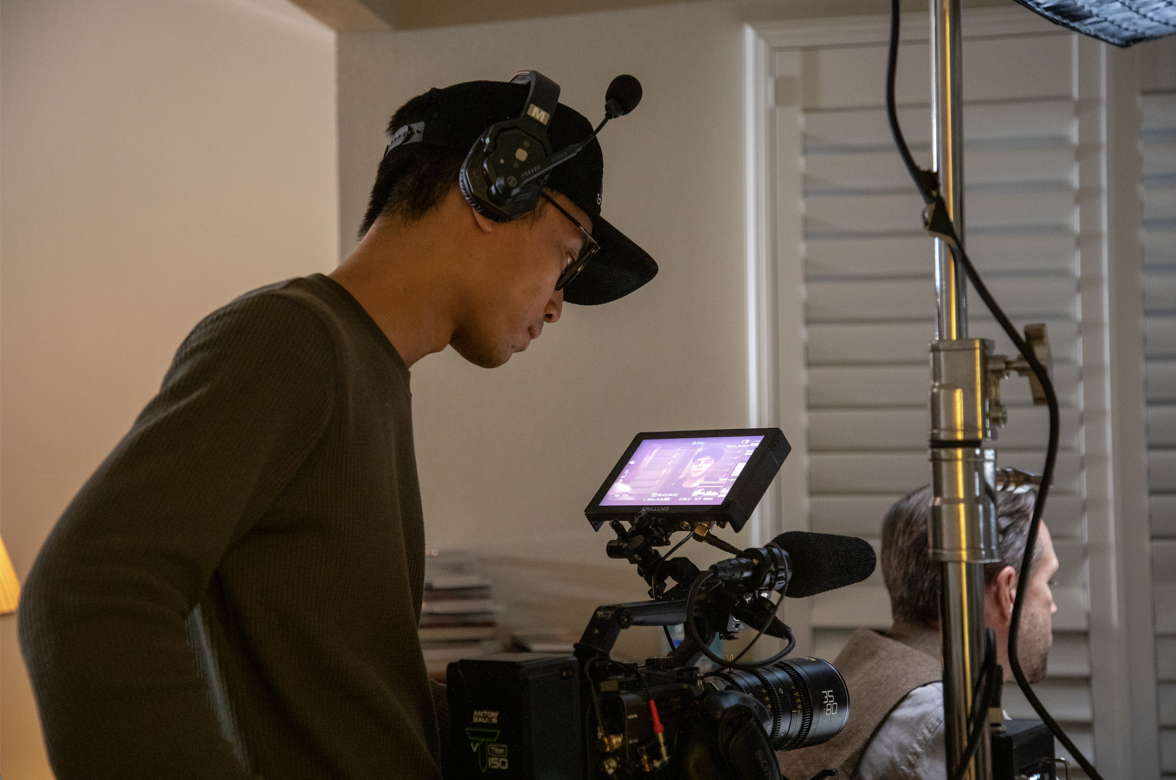 A man wearing a black cap, glasses, and a black shirt is operating a professional camera with a mounted monitor, during filming. 