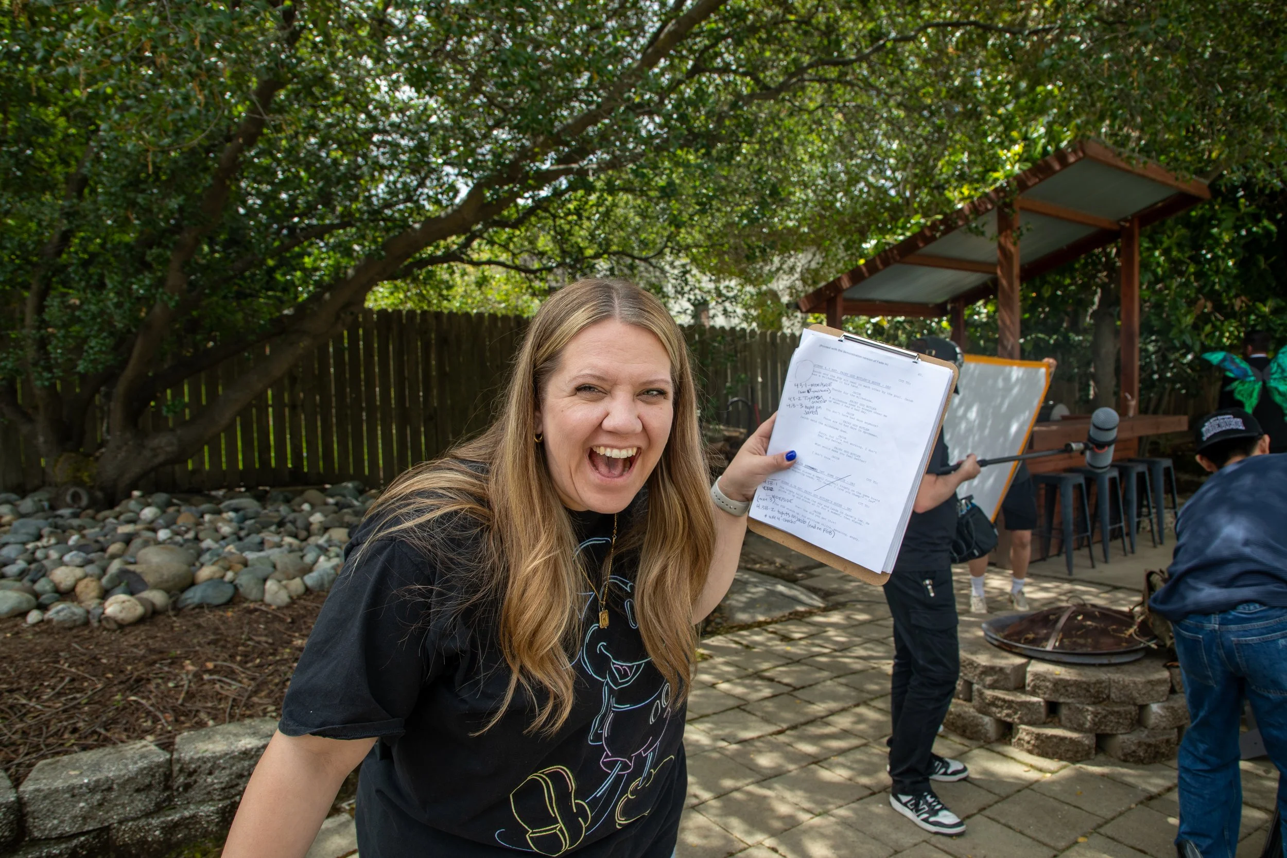 The producer with long blonde hair is smiling and holding a clipboard outdoors.