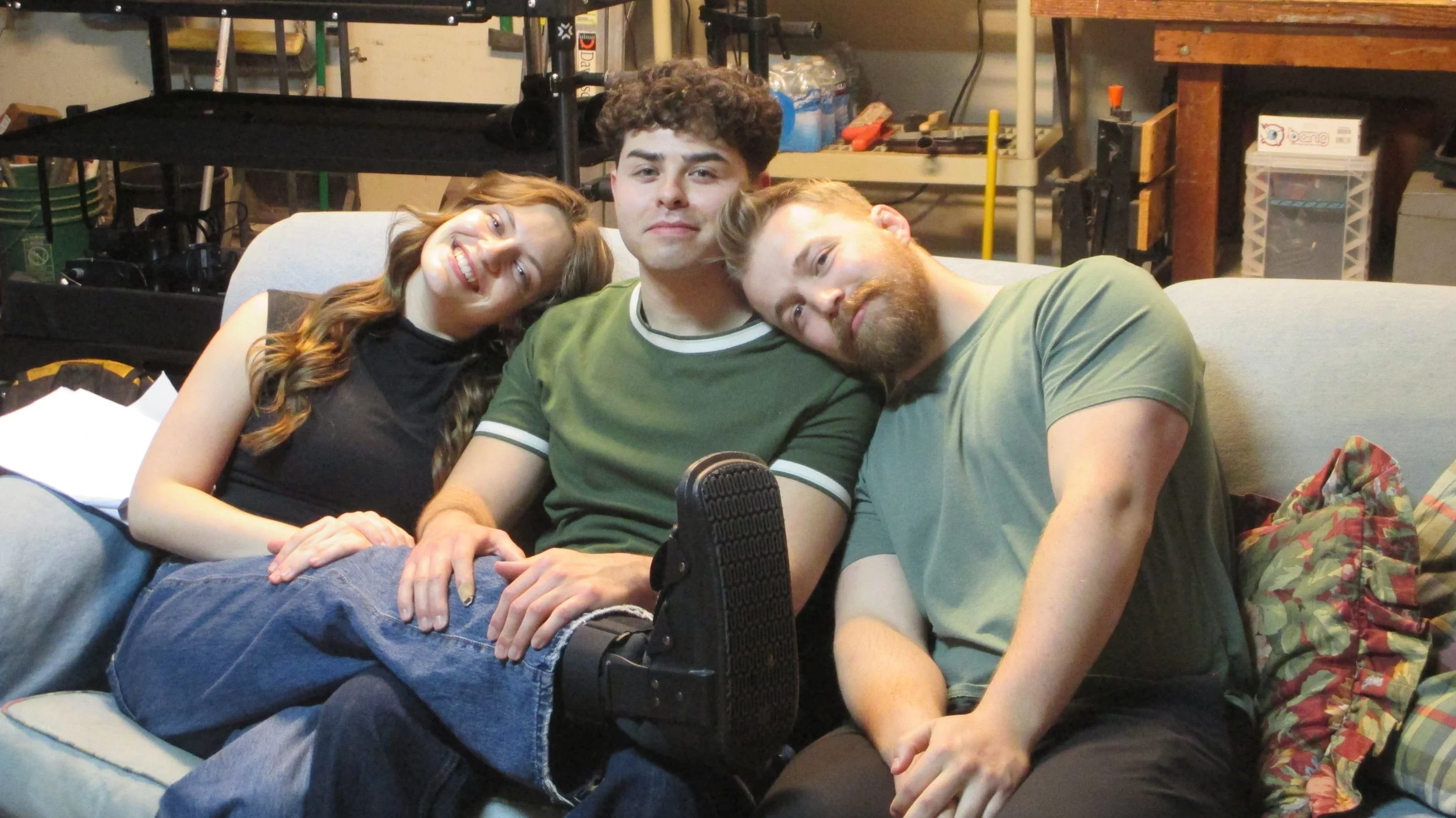 Three friends sitting closely together on a couch in a room with shelves and water bottles in the background, smiling and relaxed.