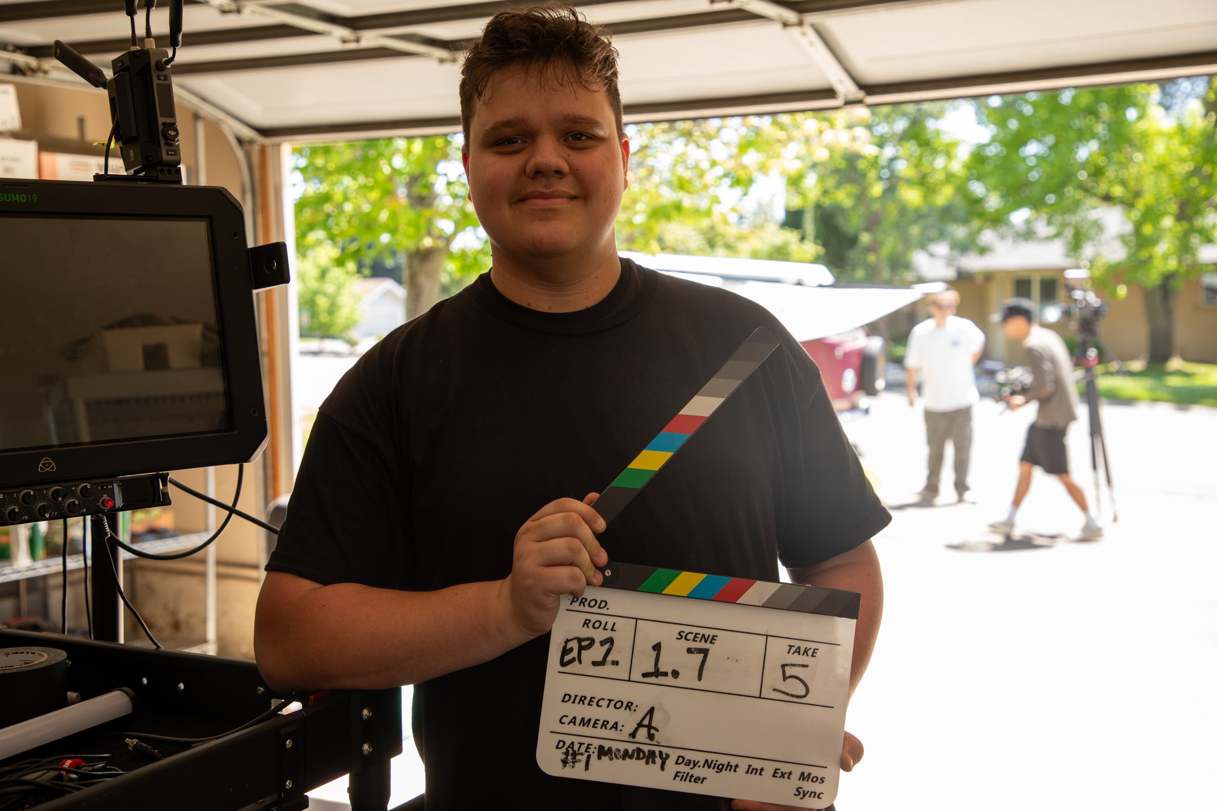 Young man holding a film slate in front of film equipment, standing outdoors with trees and a few people in the background.