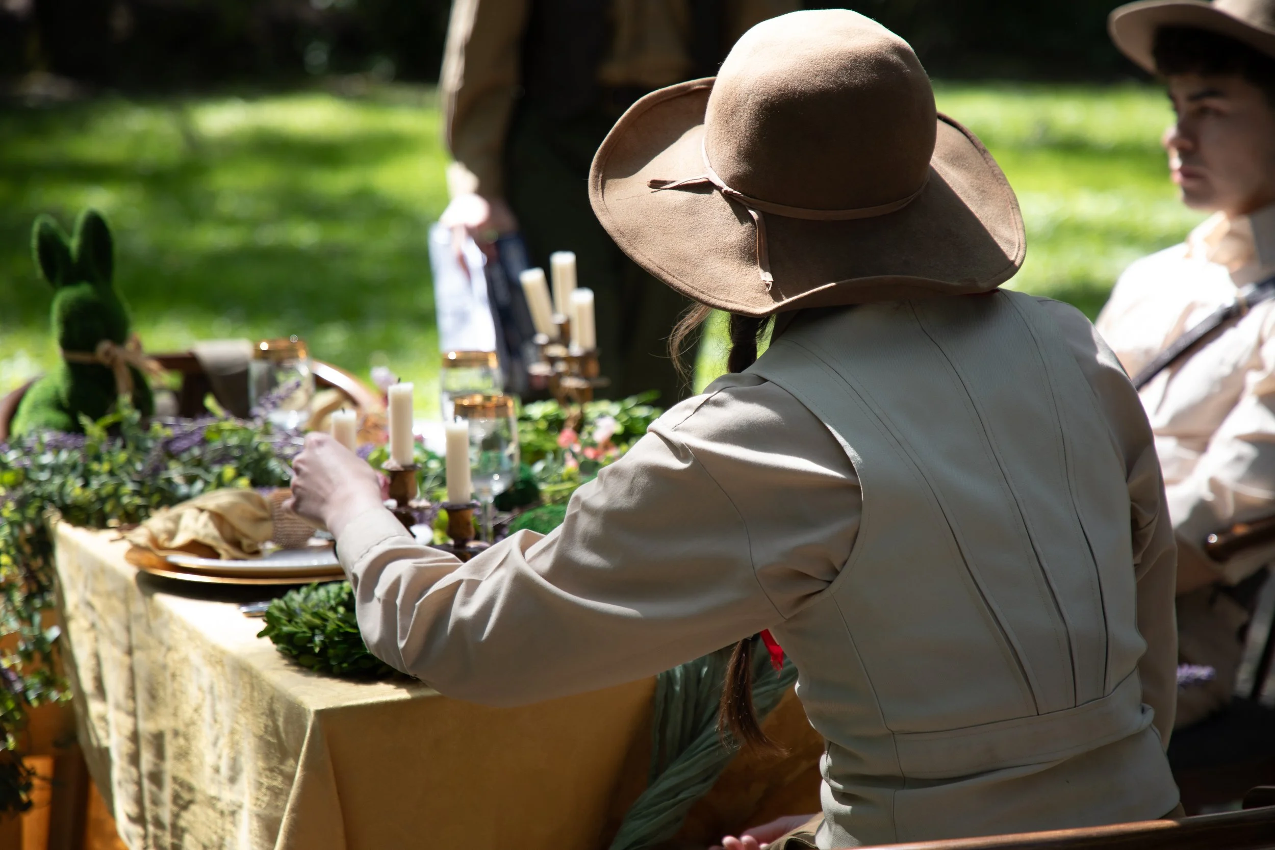 A woman wearing a large beige hat sits at an outdoor table decorated with candles and greenery, with a person in the background. The scene is set in a lush, green park or garden.