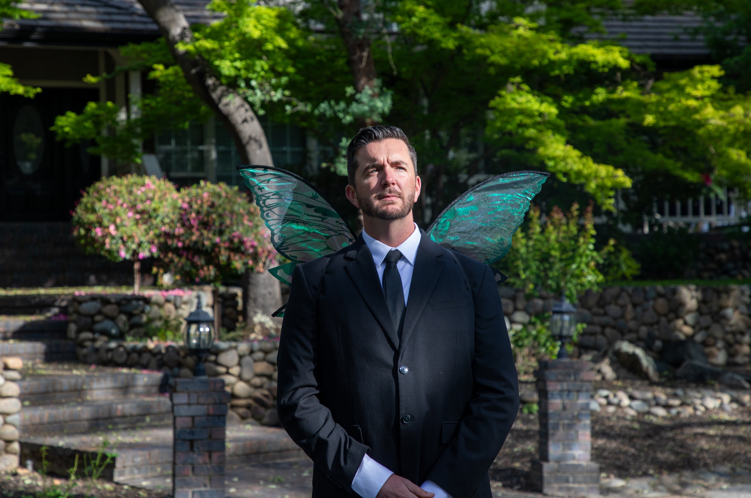 A man wearing a black suit with fairy wings standing outdoors in front of greenery and flowering bushes.